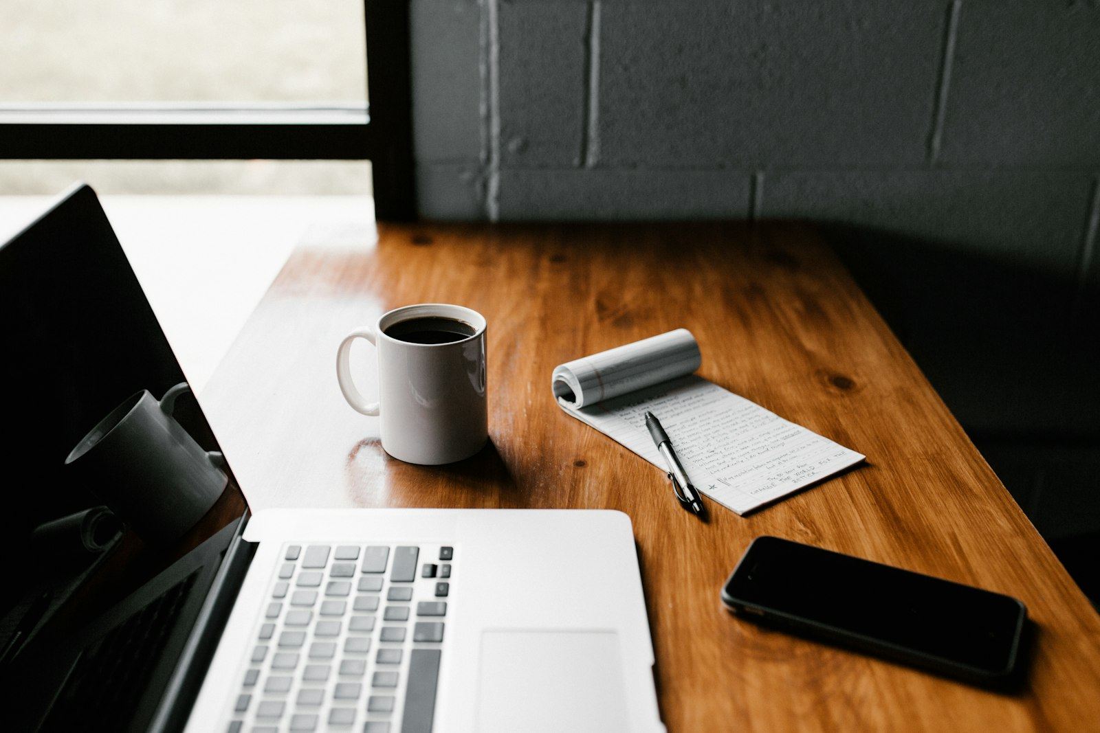 A laptop, a mug and a handwritten notebook on a wooden desk, seen from above.